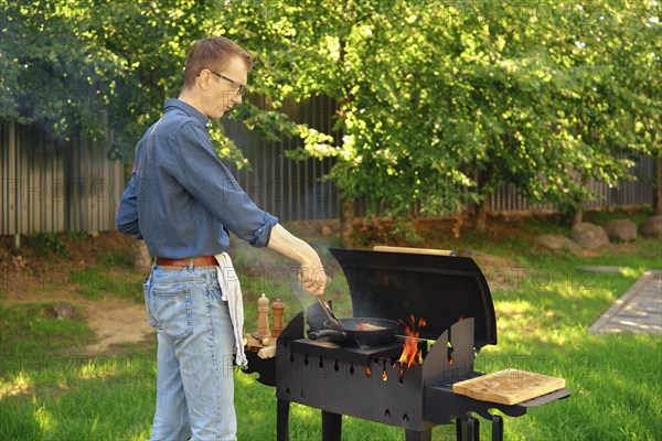 Middle-aged man puts a steak on a frying pan with kitchen tongs while making food at backyard