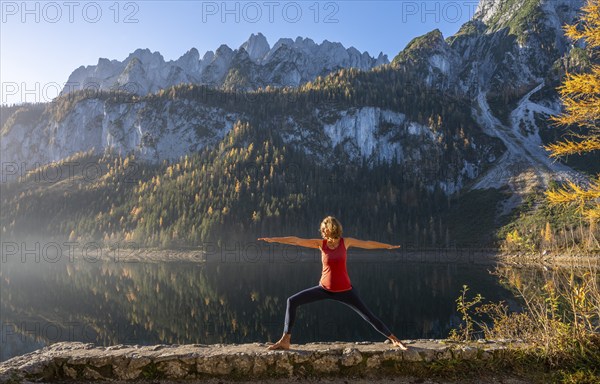 A woman doing yoga by the lake. Warrior 2 (Virabhadrasana) . Vorderer Gosausee in autumn with a