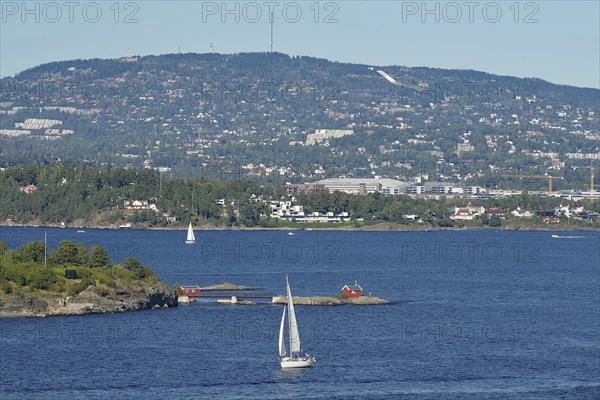 A sailboat sails on a calm sea in front of a hilly landscape with scattered houses and clear blue