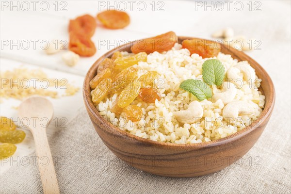 Bulgur porridge with dried apricots, raisins and cashew in wooden bowl on a white wooden background
