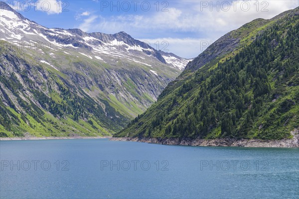 A peaceful mountain lake surrounded by green mountains, snow-capped peaks and a clear blue sky,