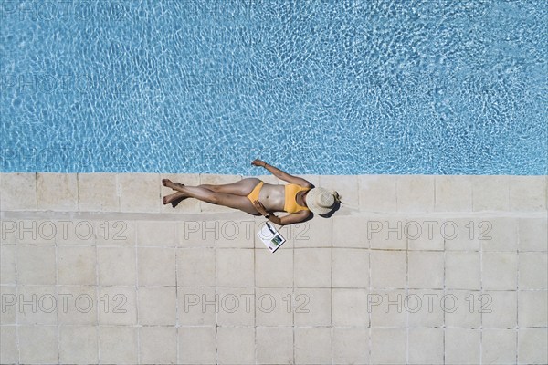 Young woman lying in the sun at the edge of a swimming pool, her face covered by a sun hat,
