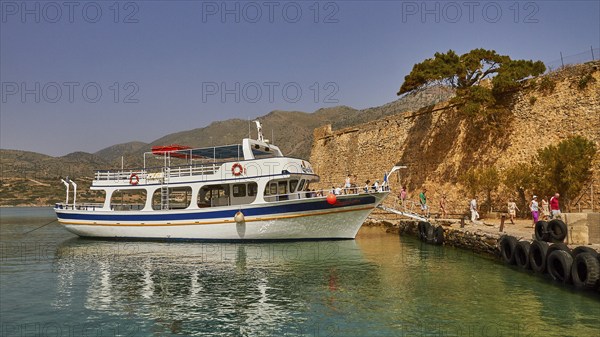 A boat moored in a quiet harbour next to a historic wall, with people on the jetty, Venetian Sea