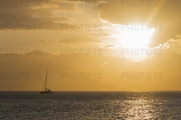 A sailboat on a calm sea at sunset with a golden sky and mountains in the background, Agia Kyriaki,