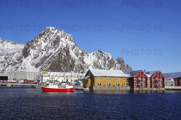 Colourful houses and a red fishing boat in the harbour in front of snow-covered mountains under a