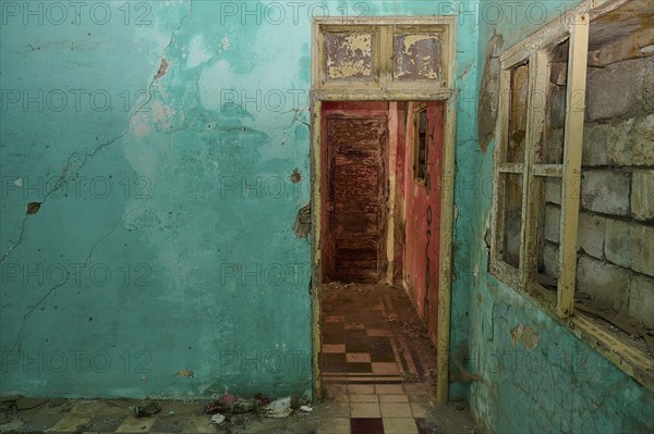 Ruined room with bright blue wall colour and destroyed door, window and a badly damaged floor,