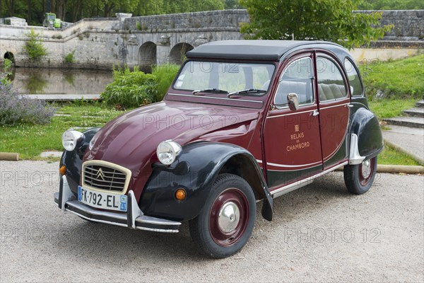 Retro Citroën 2CV vintage car in red and black, parked on a road in front of a stone bridge in a