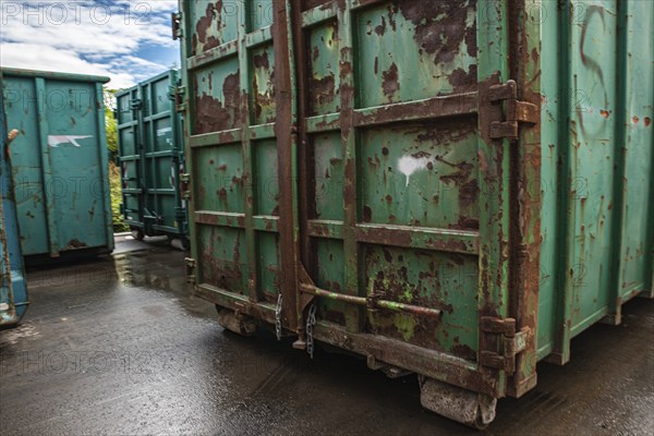 Large green containers with rust stains stand on a rain-soaked industrial site