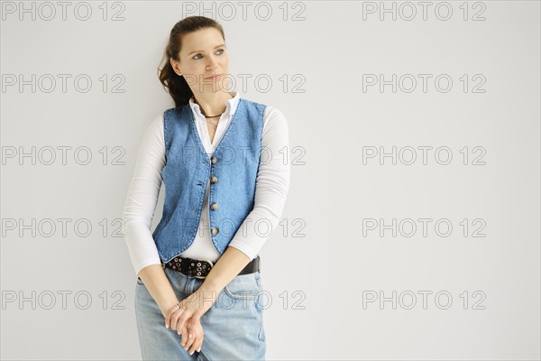 Portrait of relaxed woman in denim vest, jeans and cotton shirt leaning against white wall