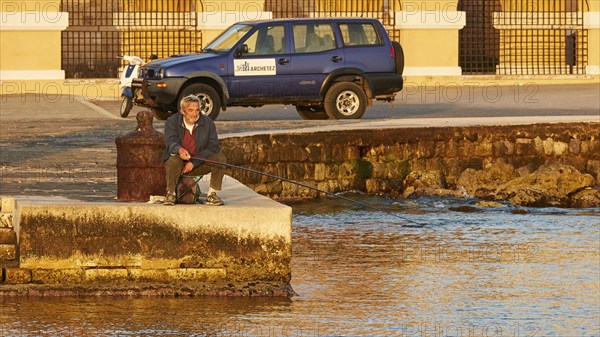 A man relaxes while fishing by the sea next to a parked off-road vehicle, Mandraki Oat, harbour