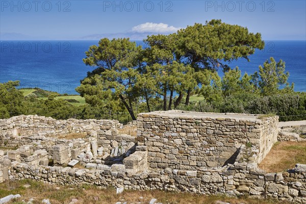 Ancient wall remains near a coast, surrounded by trees and a clear blue sky, the sea in the
