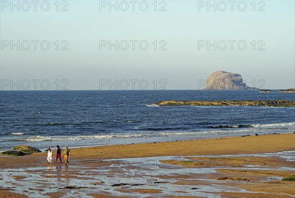 Walkers on the beach with rocks and waves in the background under a clear sky, Bass Rock, North