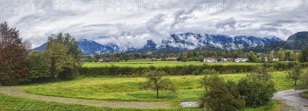 Wide landscape with green meadows, trees and a village in front of a mountain range under a cloudy