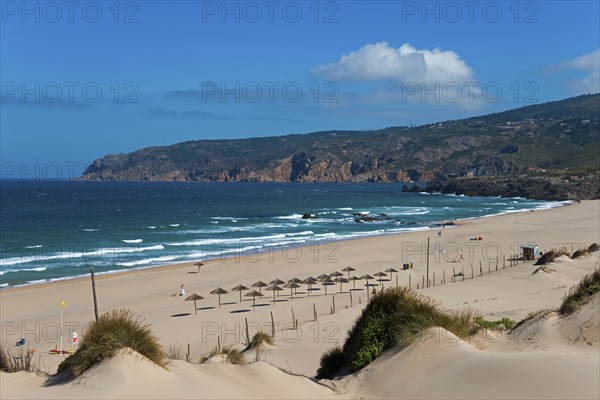 Beach with sand dunes and sunshades, behind them the sea and rocks, partly cloudy sky, moving dune,