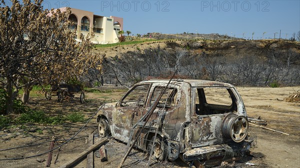Burnt car in a landscape destroyed by a fire, buildings and dry trees in the background, forest