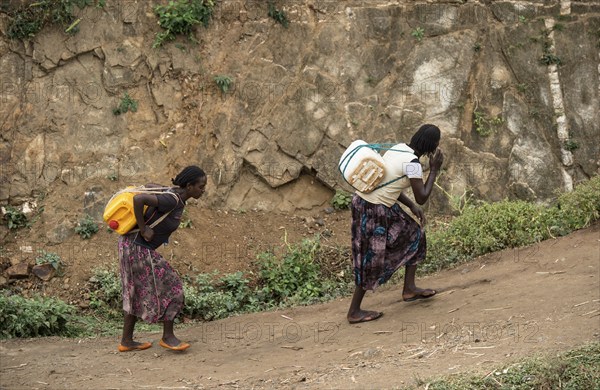 Two woman carry heavy water canisters on their backs from the water point to the village, near the