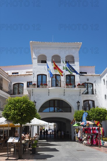 City gate of a whitewashed building on a square with flags and shops under a clear blue sky, town