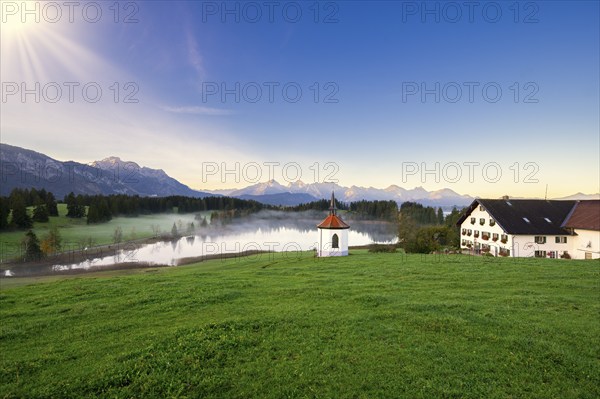 Farmhouse next to a chapel on a lake with a view of the Alps at sunrise, surrounded by green
