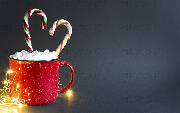 Red mug with marshmallows and caramel candy cane in the shape of a heart and lights of garlands on