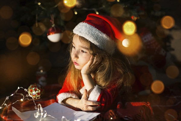 Little girl in a Santa hat and red dress under Christmas tree is dreaming, waiting for the holiday,