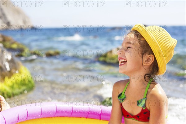 Girl in yellow straw hat plays in outdoor near sea, in water with a bucket in an inflatable pool on