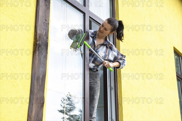 A woman manually washes the window of the house with a rag with a spray cleaner and a mop outside.