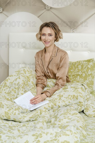 Happy woman sits in bed under the blanket, she puts her hands on a book