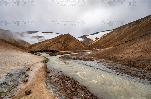 Bridge over steaming stream between colourful rhyolite mountains with snowfields, Hveradalir