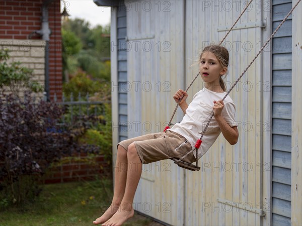 Boy swinging while sitting on children's swing at shed and garden gate, Germany, Europe