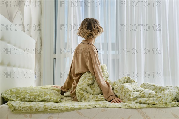 Back view of young woman stretching arms sitting on the bed at home bedroom looking to the window
