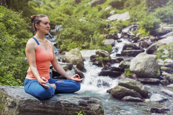 Woman in Hatha yoga asana Padmasana outdoors at tropical waterfall