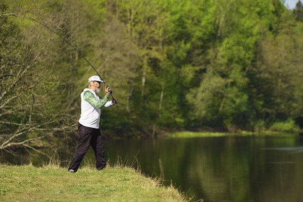 Fisherman with a spinning rod catching fish on a river at sunny summer day with green trees at