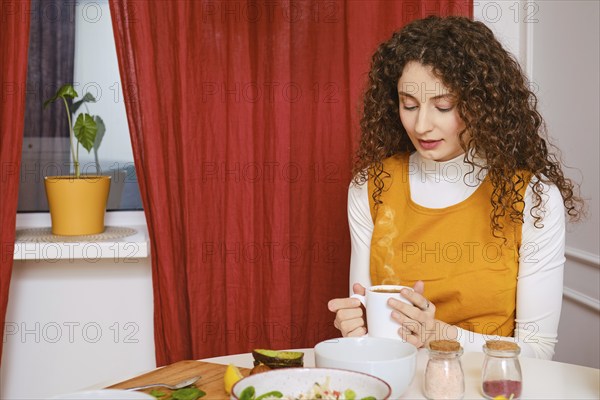 Peaceful young woman holds cup of coffee with both hands while having breakfast