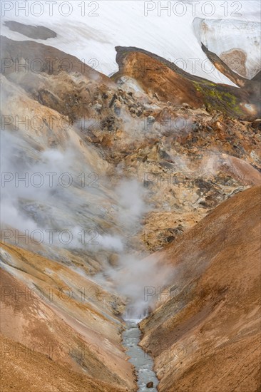 Steaming stream between colourful rhyolite mountains and snowfields, Hveradalir geothermal area,