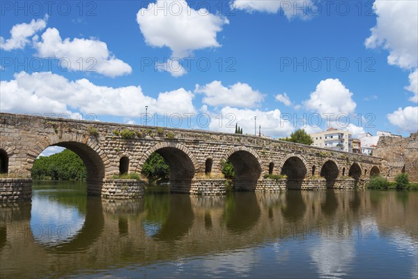 Ancient stone bridge with reflections in the river water, under blue sky with some clouds, Roman