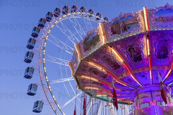 A brightly lit carousel in front of a large Ferris wheel at dusk at a funfair, Wellenflug,