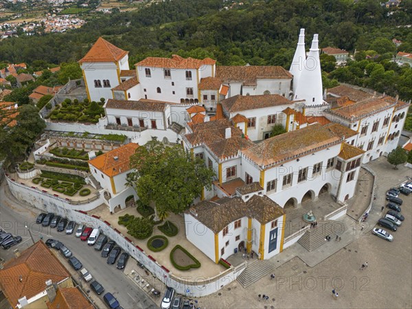 Overview of a historic castle with white towers and orange-red roofs, surrounded by a city, aerial