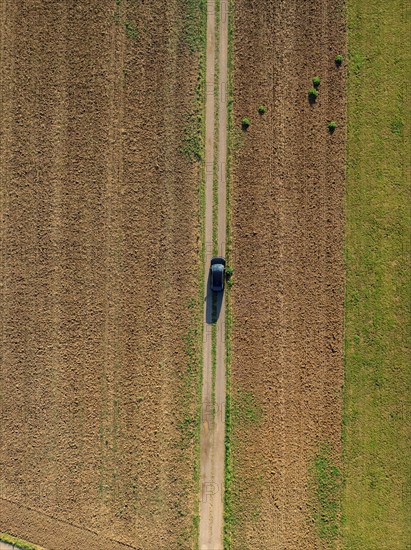 Aerial view of a car on a narrow country lane between green and brown fields on a clear day, car