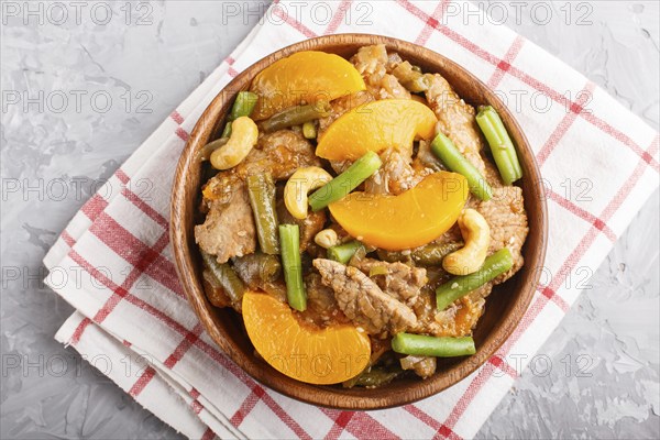 Fried pork with peaches, cashew and green beans in a wooden bowl on a gray concrete background. Top