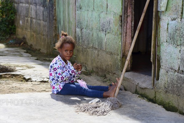 A young girl plays on the ground next to a broom in front of a house in the countryside, between