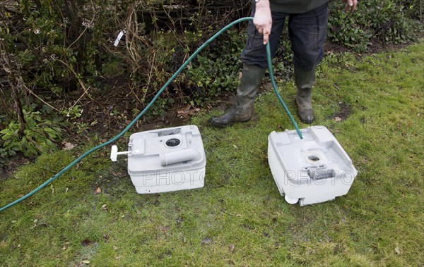 Woman using garden hose water pipe to wash clean camping toilet container cartridge, UK