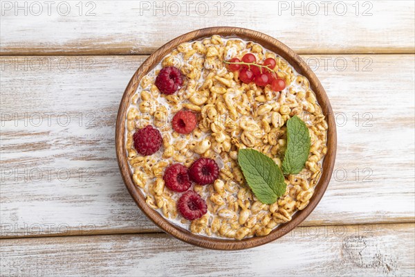 Wheat flakes porridge with milk, raspberry and currant in wooden bowl on white wooden background.
