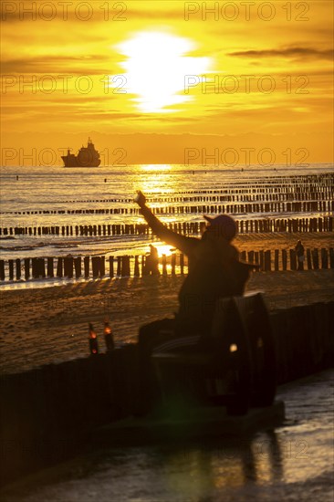 Sunset on the beach of Zoutelande, beach with wooden pile breakwaters, tourists, cargo ship sailing