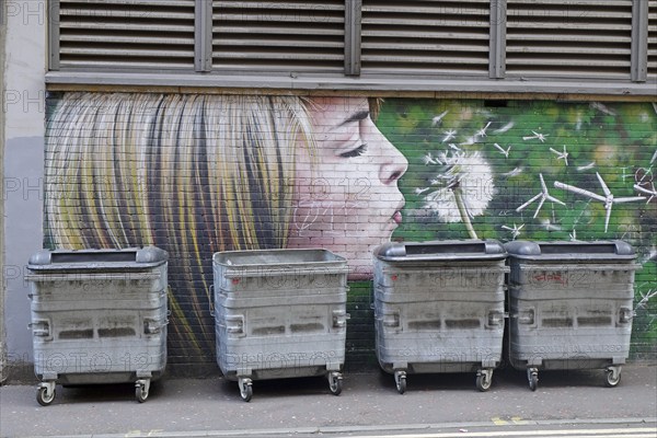 A mural shows a girl blowing a dandelion in front of rubbish containers, Mural Trail, Glasgow,