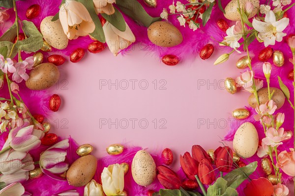 Frame of eggs and spring flowers on pink feathers with red and gold chocolate eggs on a pink