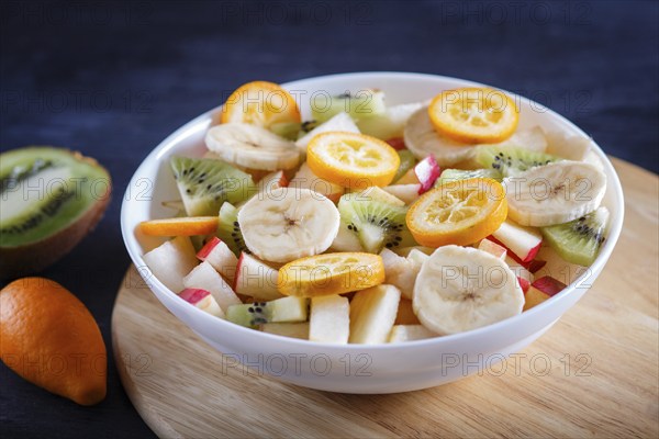 Vegetarian salad of bananas, apples, pears, kumquats and kiwi on black woodem background, close up,