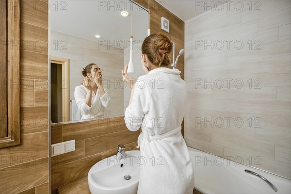 Woman thoroughly washing her face with skin cleanser while standing in a bathrobe in front of a
