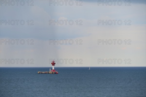 A red lighthouse stands on a calm, blue sea under a slightly cloudy sky, with a small sailing boat