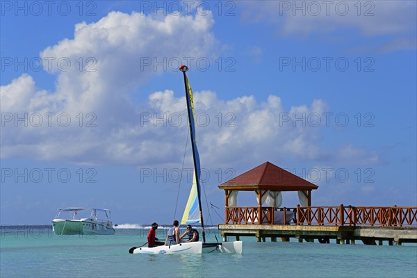 A colourful catamaran sails next to a jetty near a pavilion on the clear sea under a blue sky,