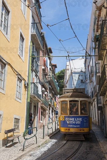 A yellow-blue tram runs through a narrow street past colourful, historic buildings on a sunny day,